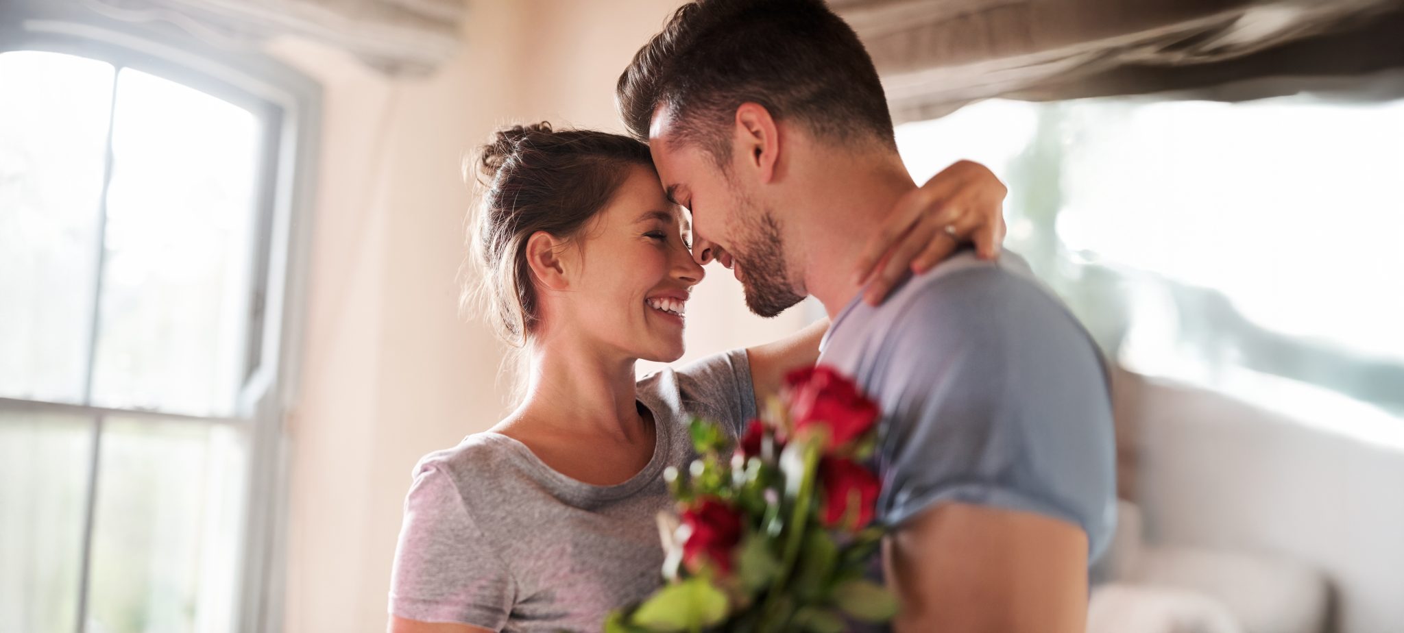 Flowers, happy couple and present in home for love, connection and bonding together with hug. Affection, man and woman with red roses for relationship, commitment and anniversary celebration in house